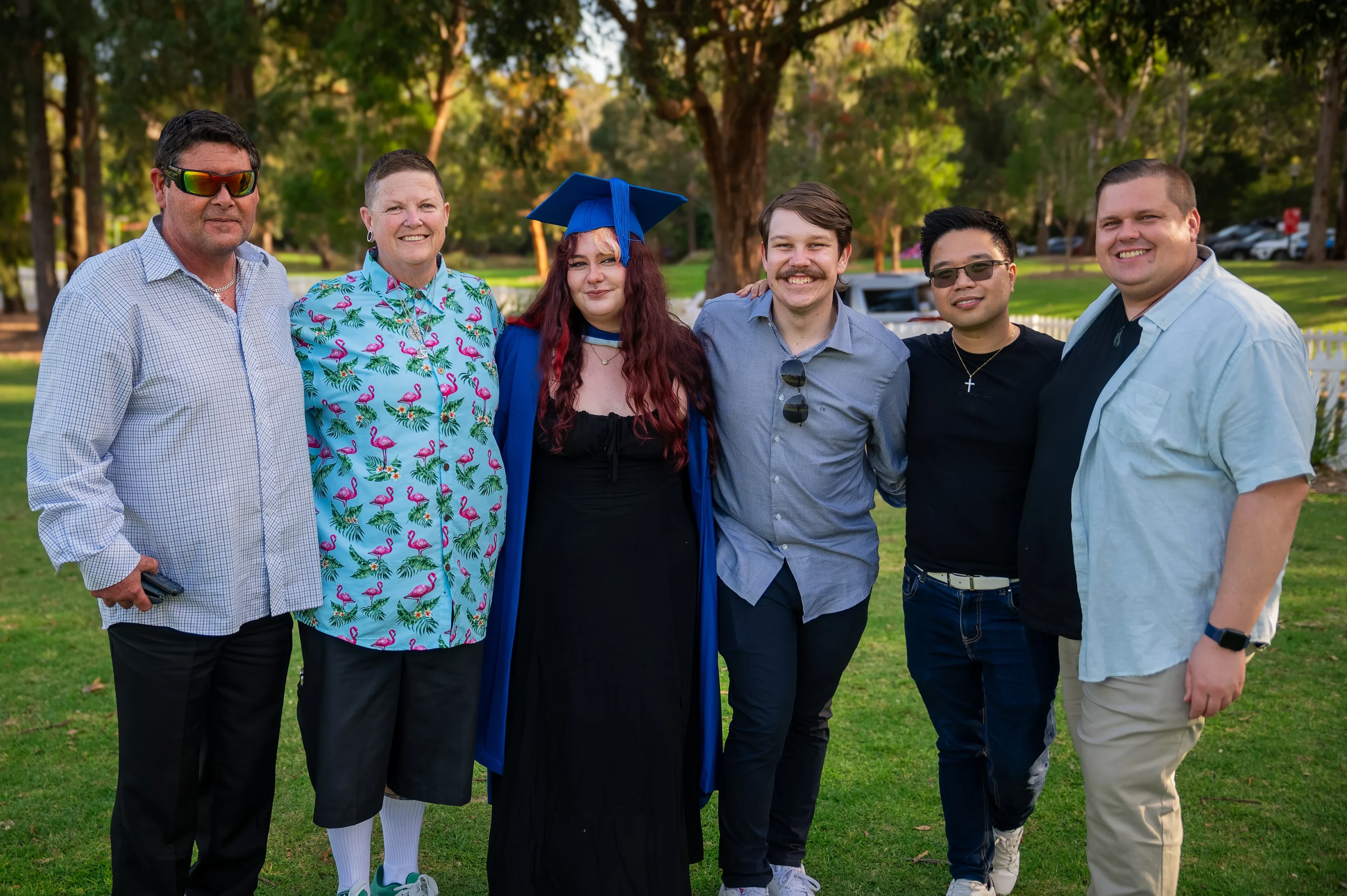 Six People On Grass; One In Blue Graduation Cap And Gown; Trees In Background.