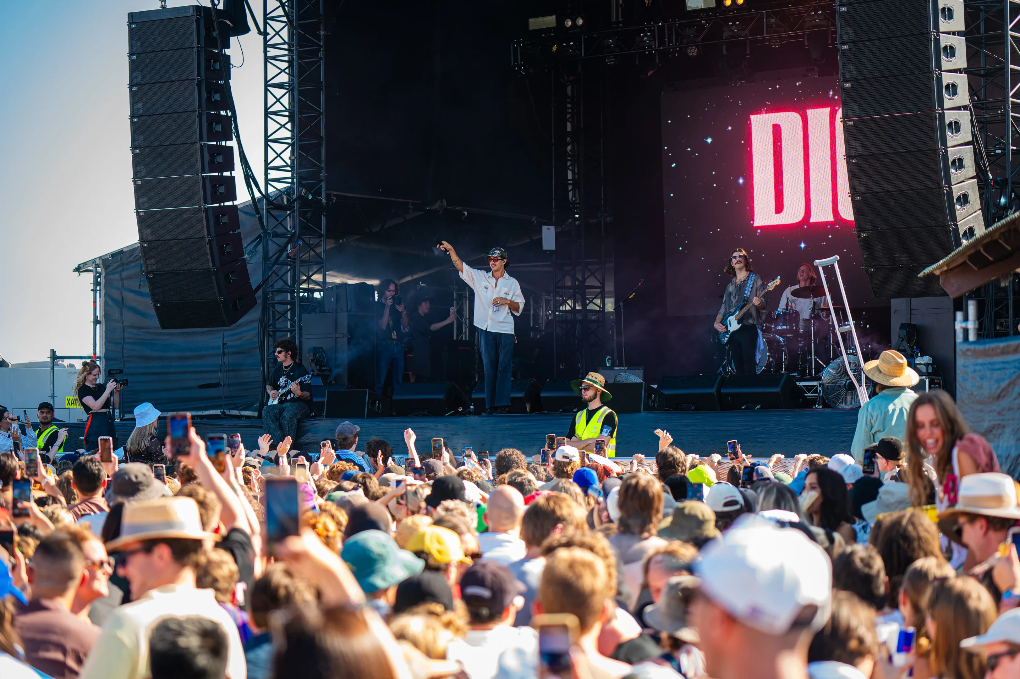 Outdoor Concert With Energetic Performer On Stage, Crowd Recording, Security Present.