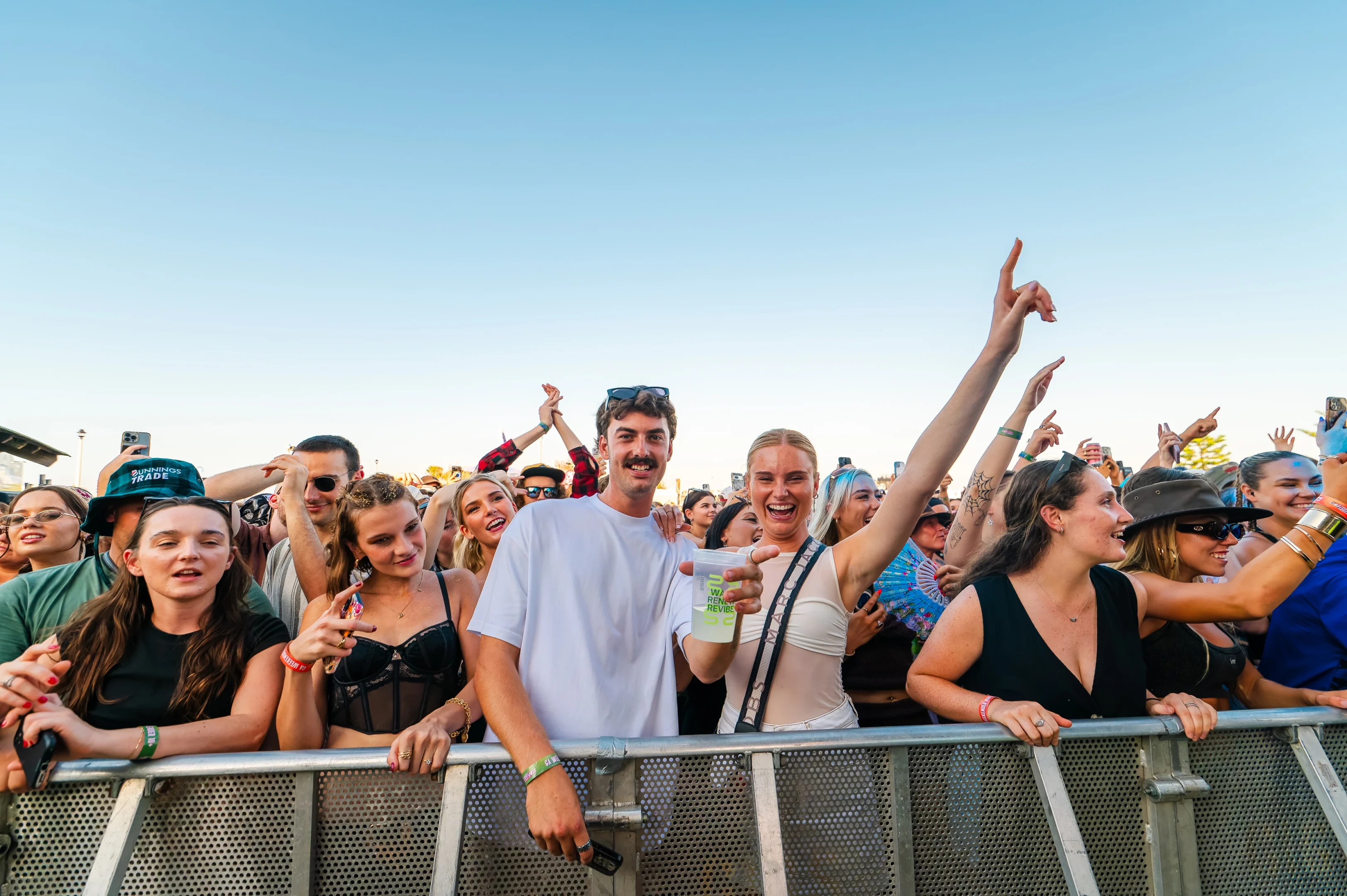 A Crowd Enjoys An Outdoor Event Near A Metal Barrier Under A Clear Blue Sky.