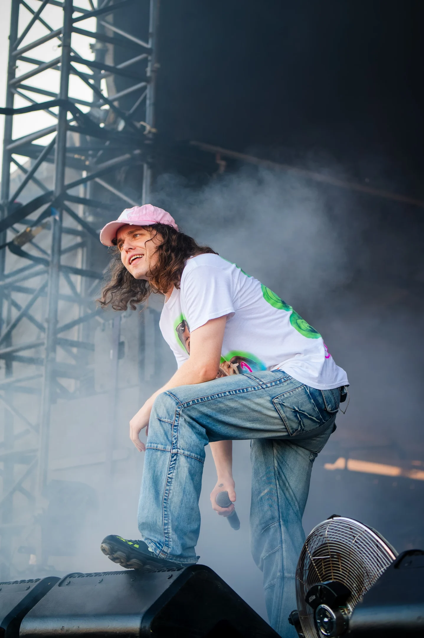 Person On Stage With Microphone, Pink Cap, White Shirt, Blue Jeans, Fog, And Visible Fan.