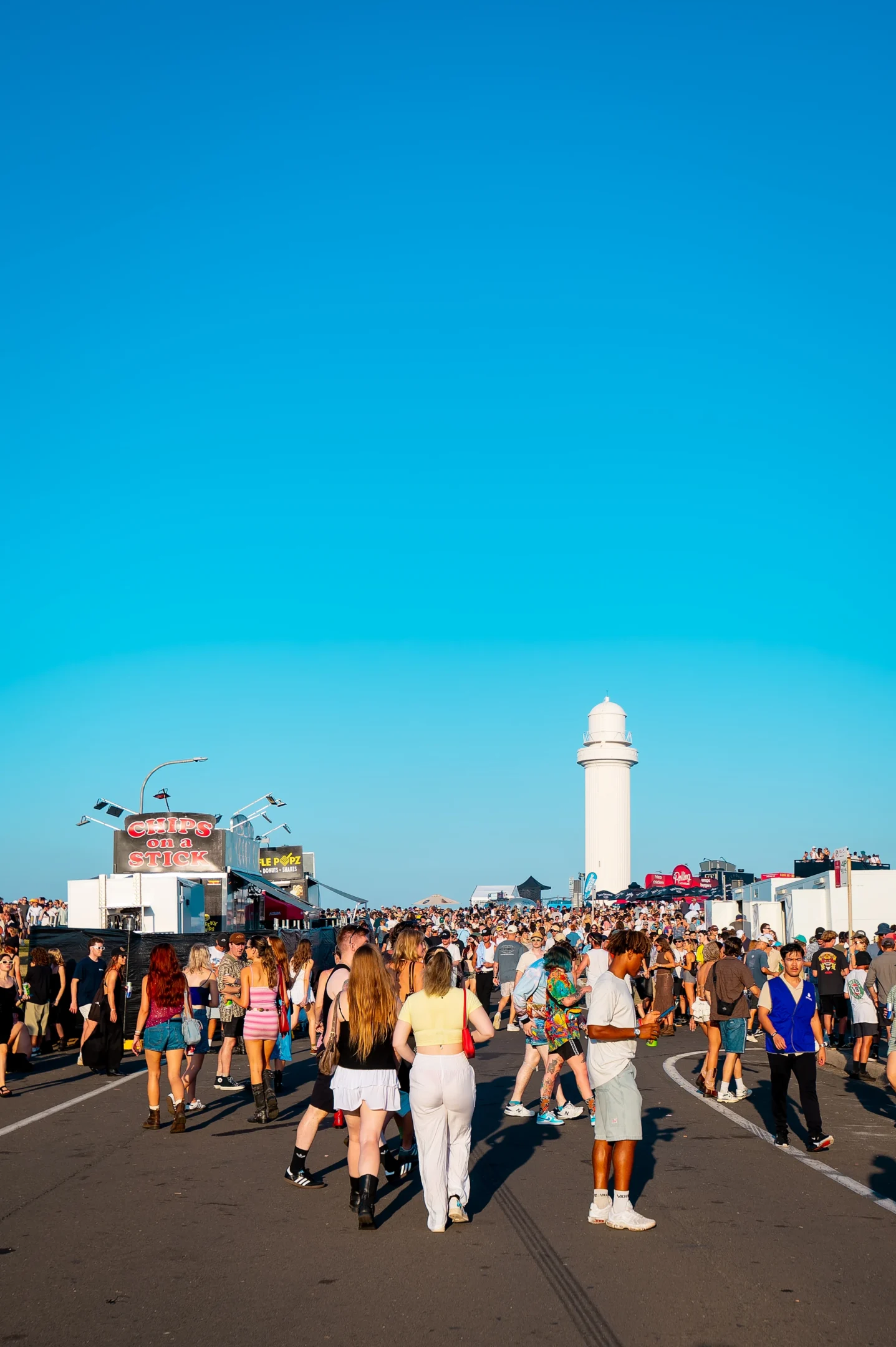 Outdoor Festival With Large Crowd, Food Stall, Lighthouse, And Clear Blue Sky.