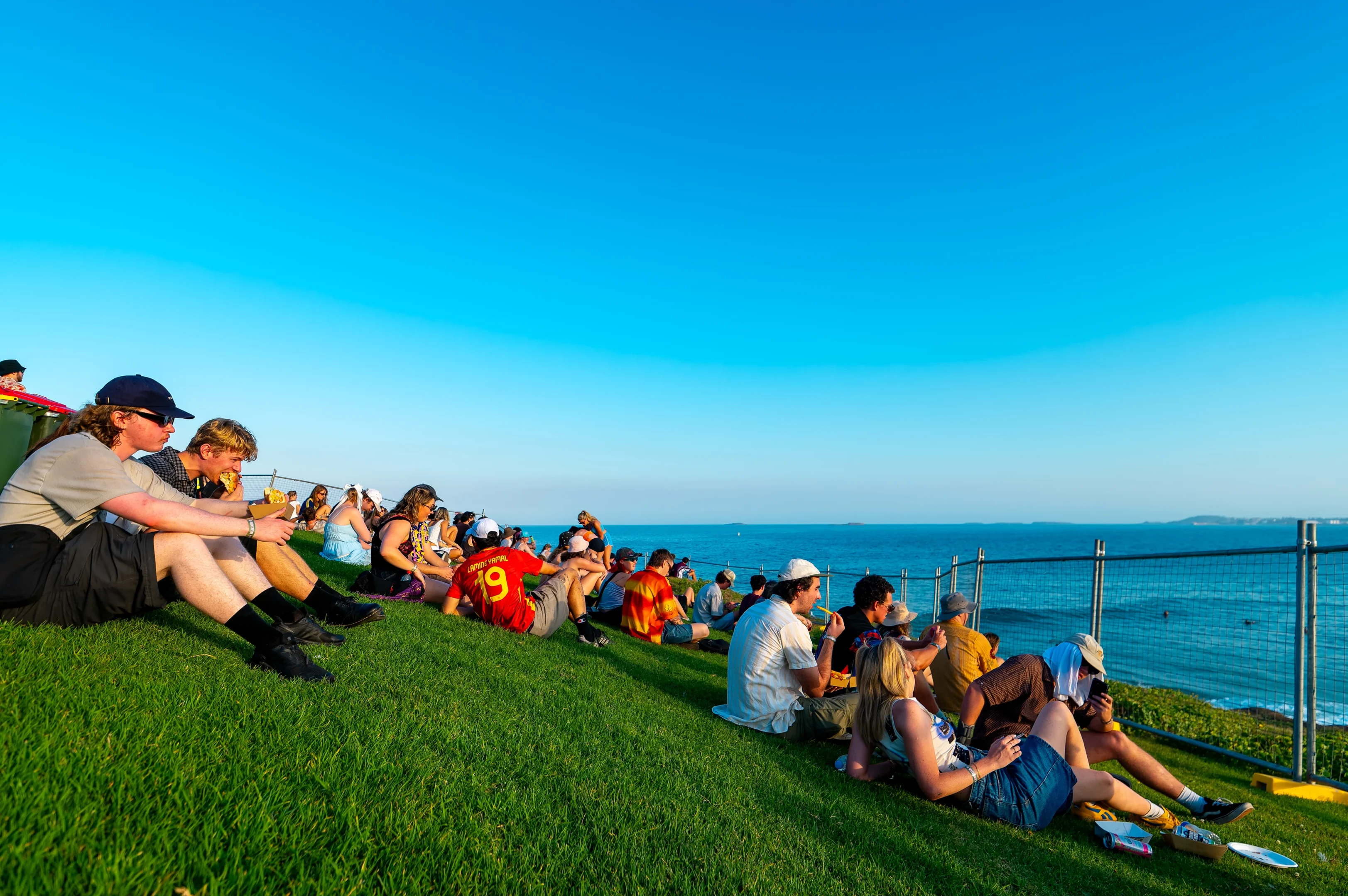 Group Seated On A Grassy Hill Overlooking Ocean, Clear Sky, Sunny Day, Some Wear Hats And Sunglasses.