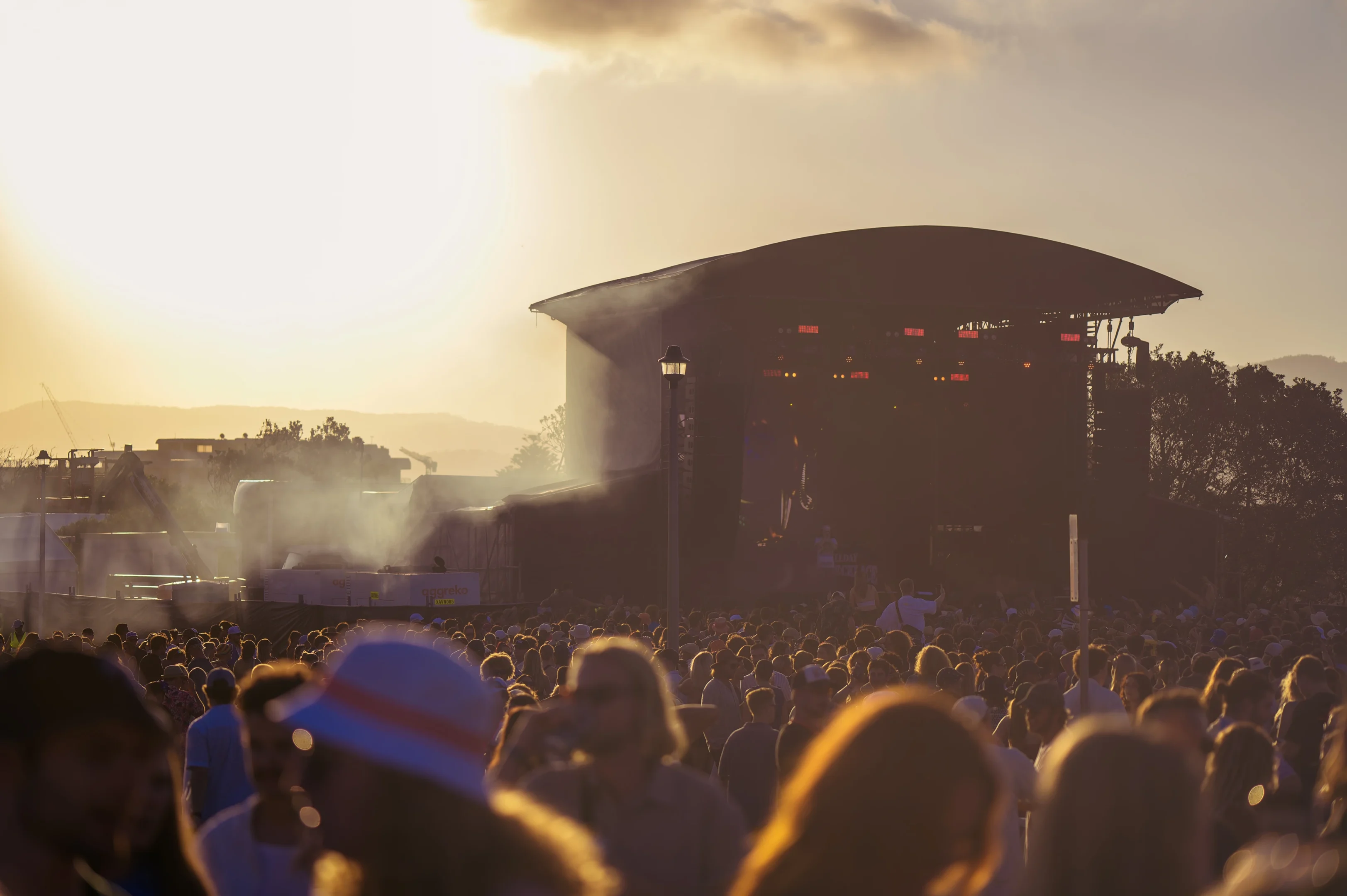 Large Outdoor Concert Crowd At Sunset, Stage Illuminated, Haze And Silhouettes Visible.