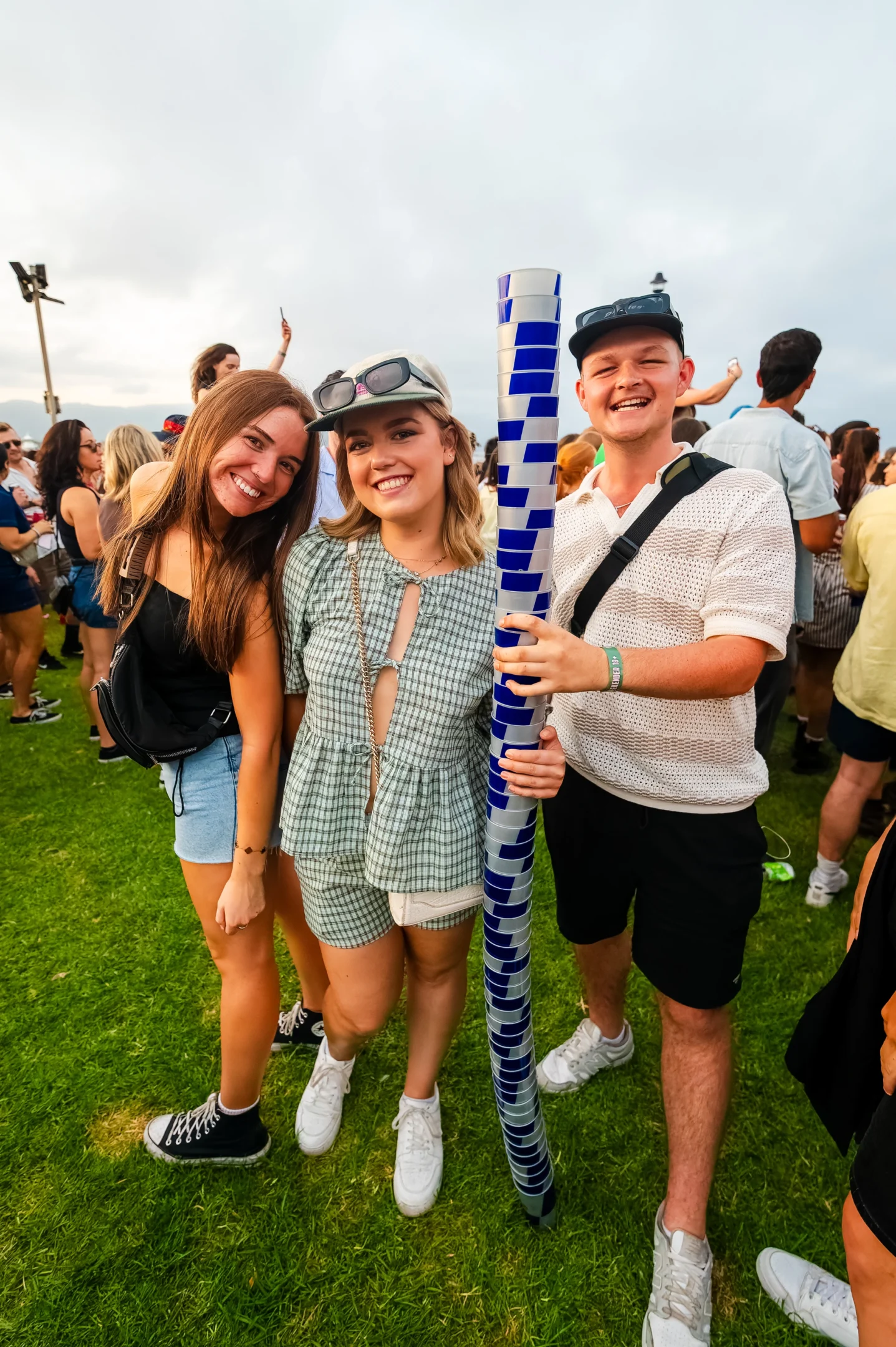 Three People Smiling On Grass At Outdoor Event; One Holds A Tall Stack Of Blue And White Cups.