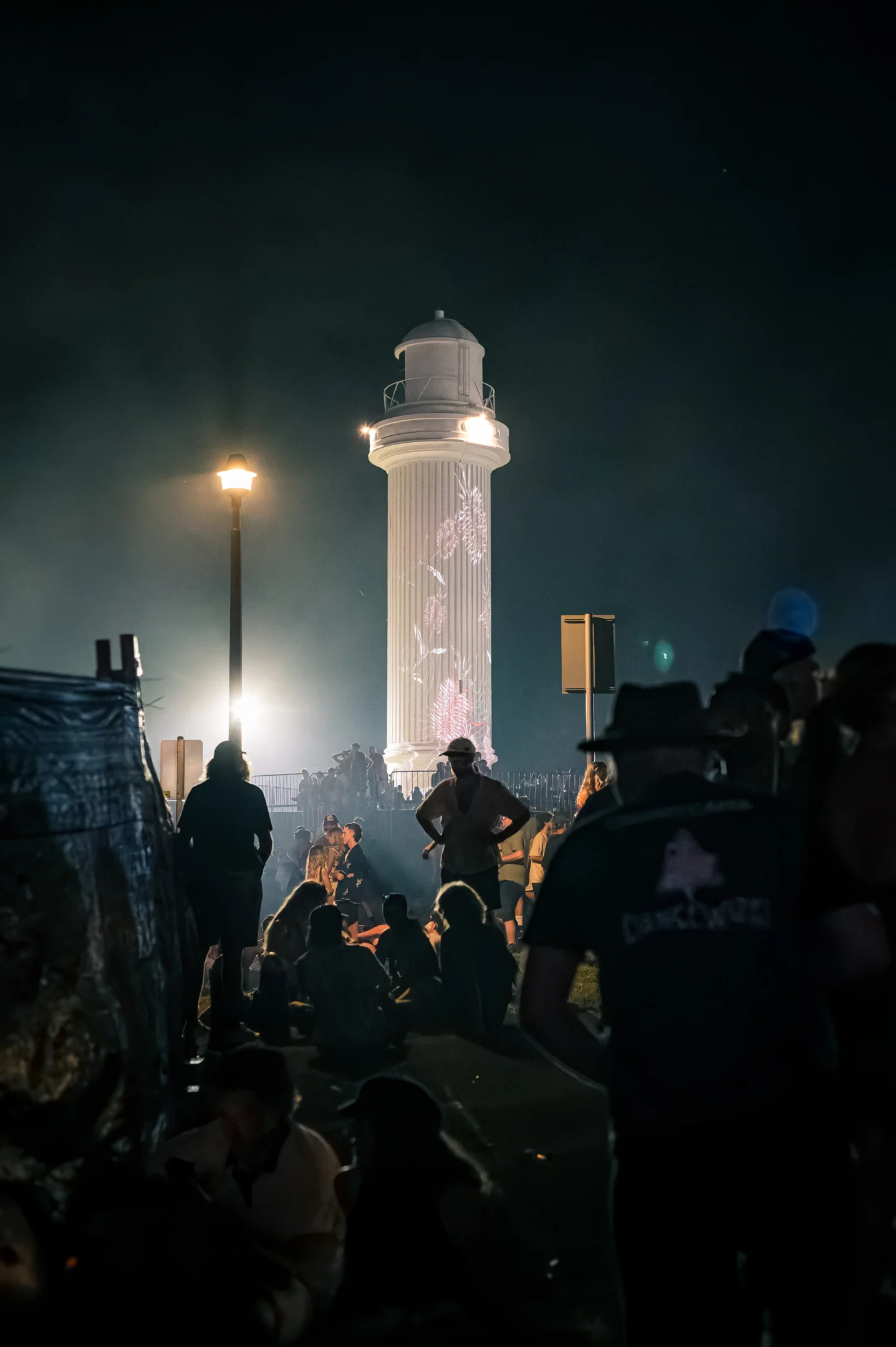 Night Gathering Near Illuminated Lighthouse With Projections, Silhouettes In Foreground.