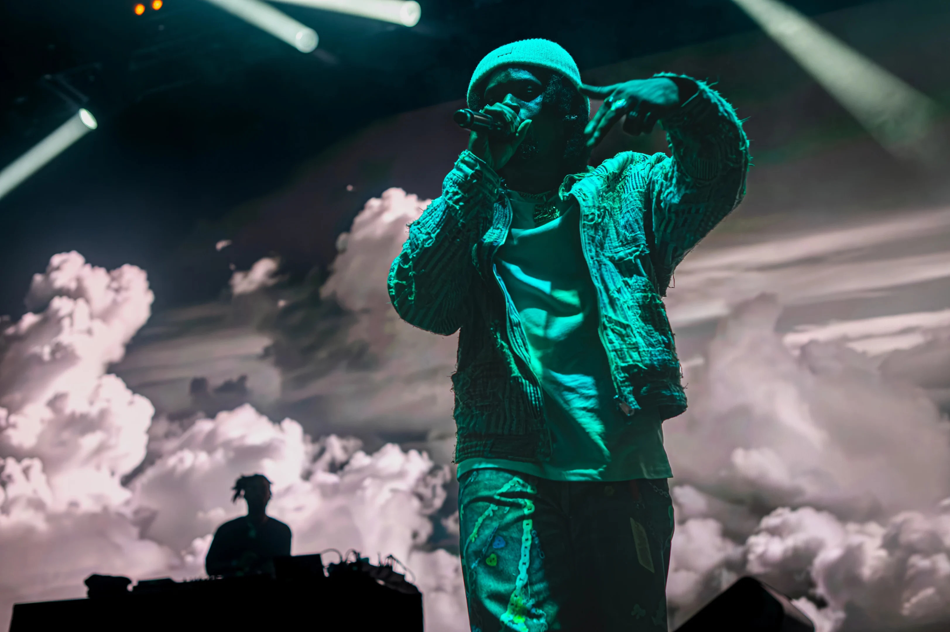 Performer Singing With A Beanie, Denim Jacket; Cloudy Background And Dj On Stage.