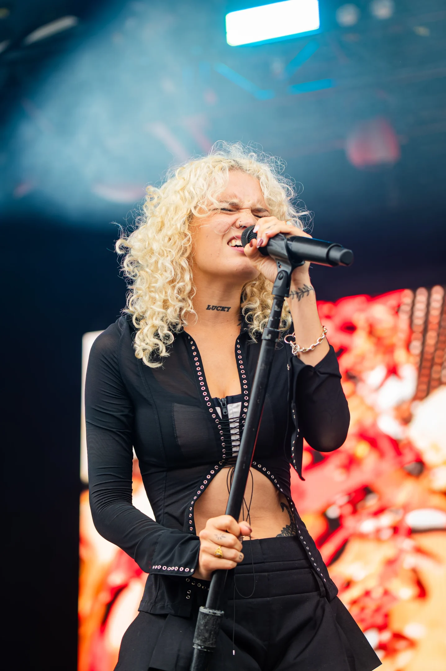 Person With Curly Hair Singing Into A Microphone On Stage With Vibrant, Colorful Background