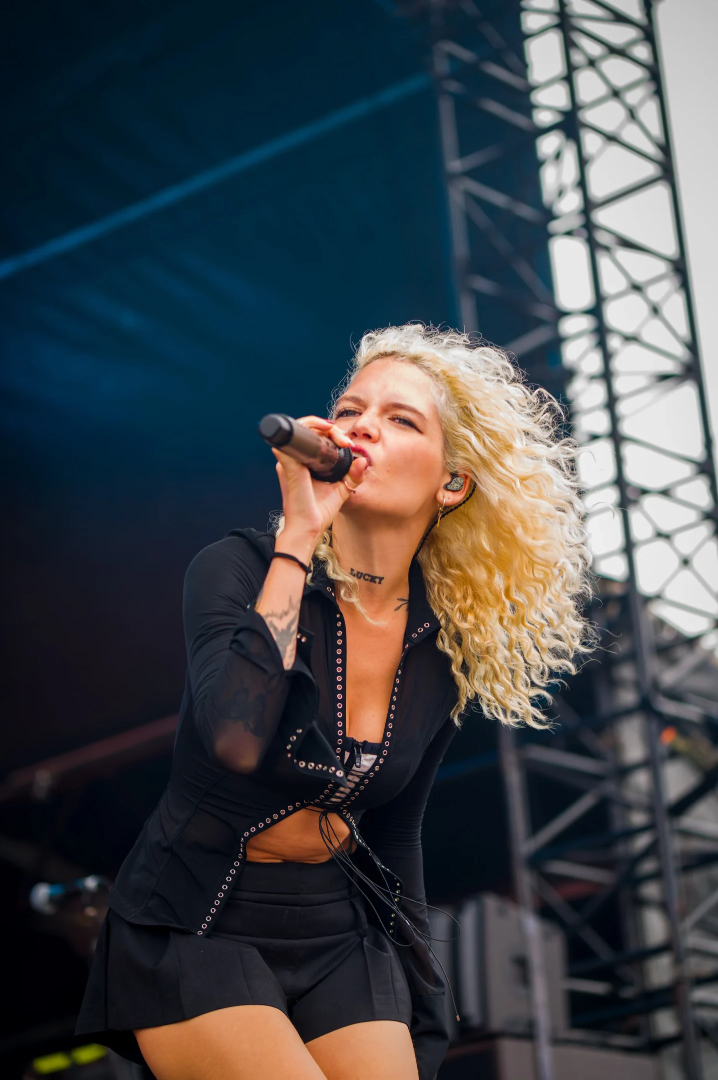 Singer With Curly Blonde Hair Performing On Stage, Wearing Black With Silver Accents.