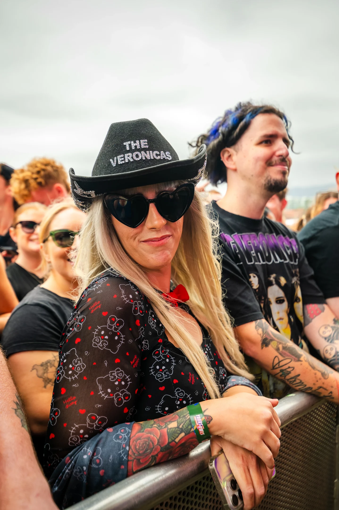 A Crowd At An Outdoor Event, Featuring A Person In A Veronicas Hat And Sunglasses By A Barrier.