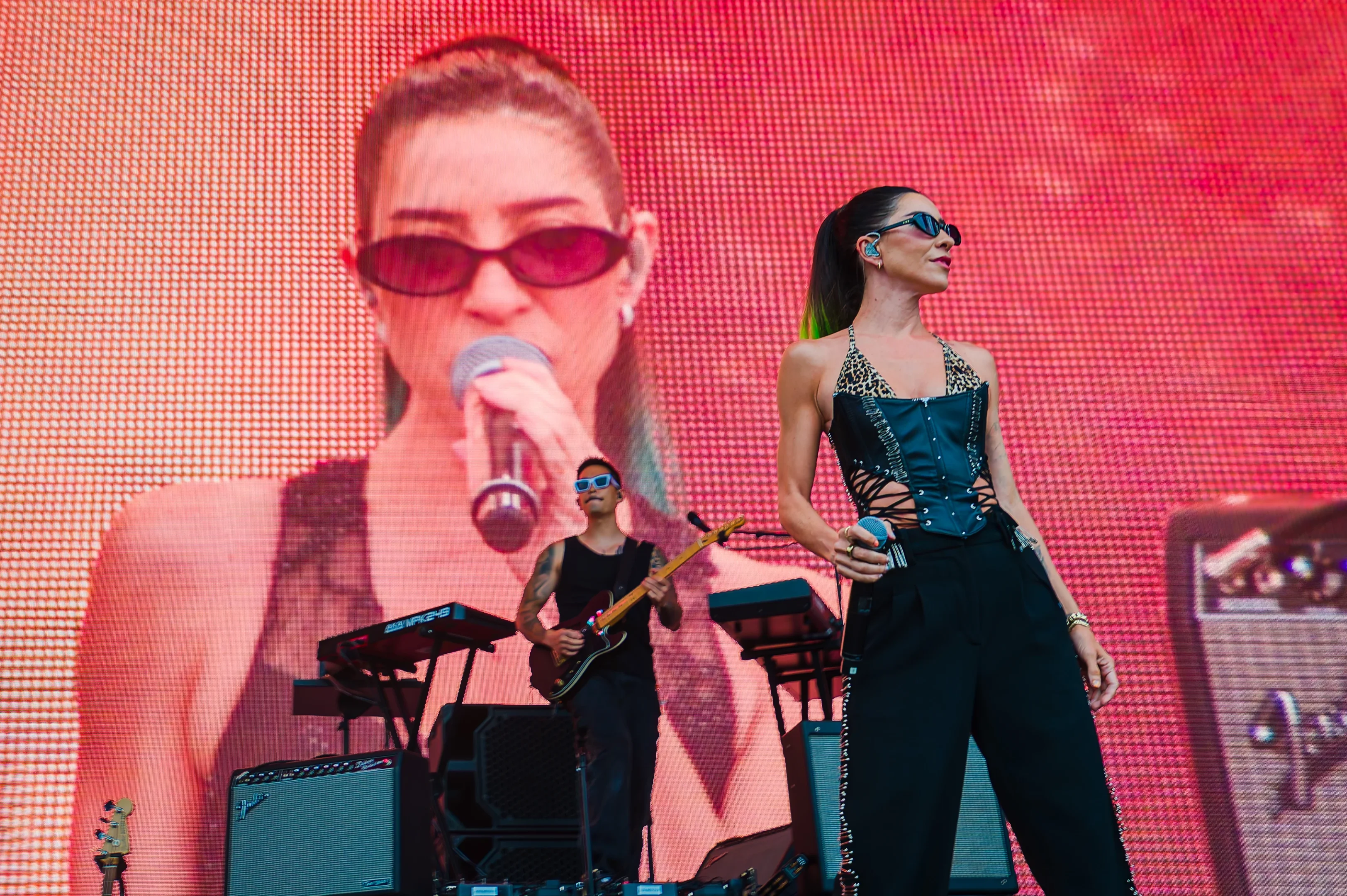 Musician Performs On Stage With Microphone, Large Screen And Band Member Playing Guitar Visible.