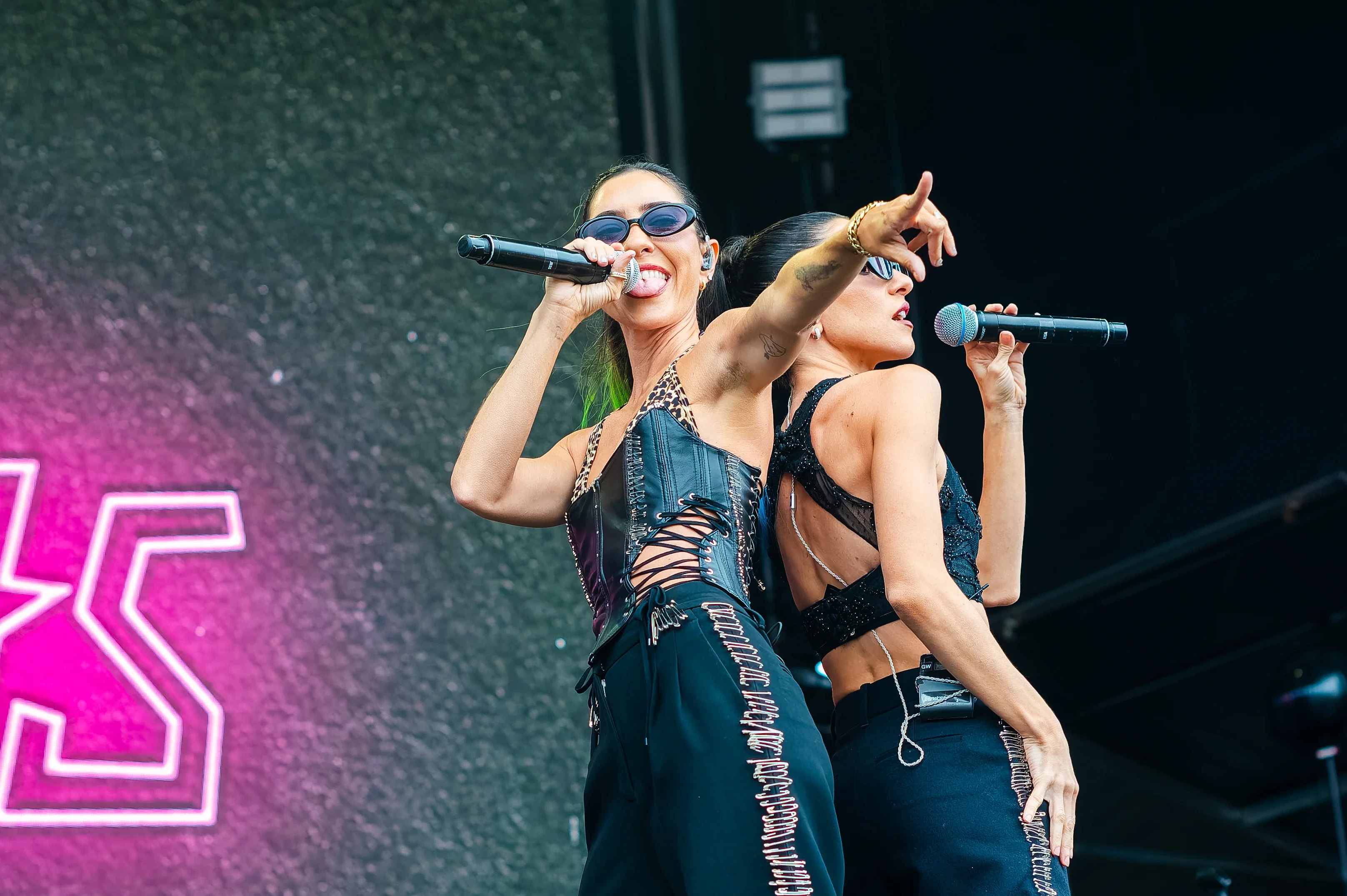 Performers On Stage With Microphones, Stylish Outfits, Neon Sign In Background.