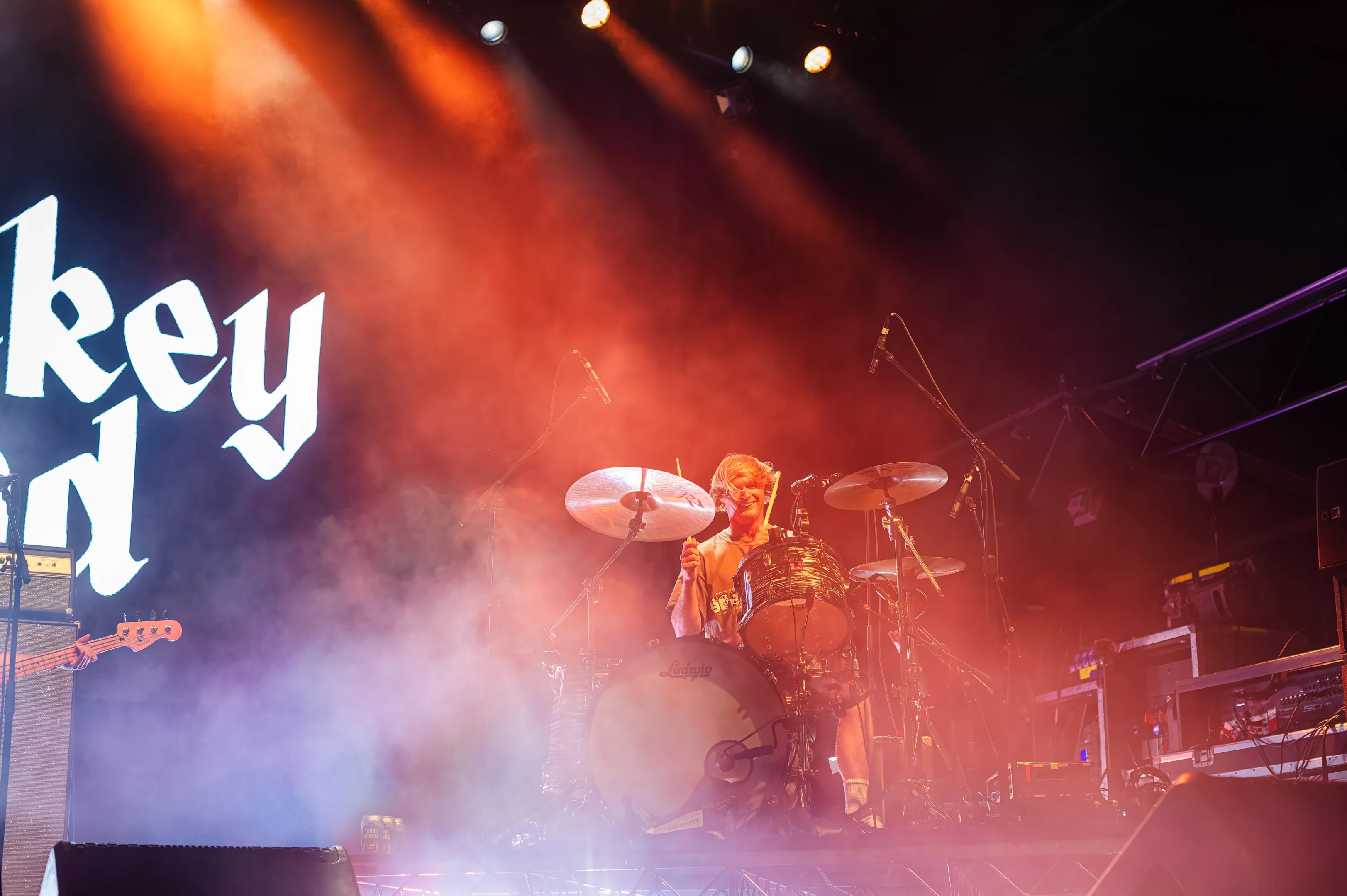 Drummer Performs On Stage With Colorful Lights, Smoke, Screen Text, And Visible Equipment.