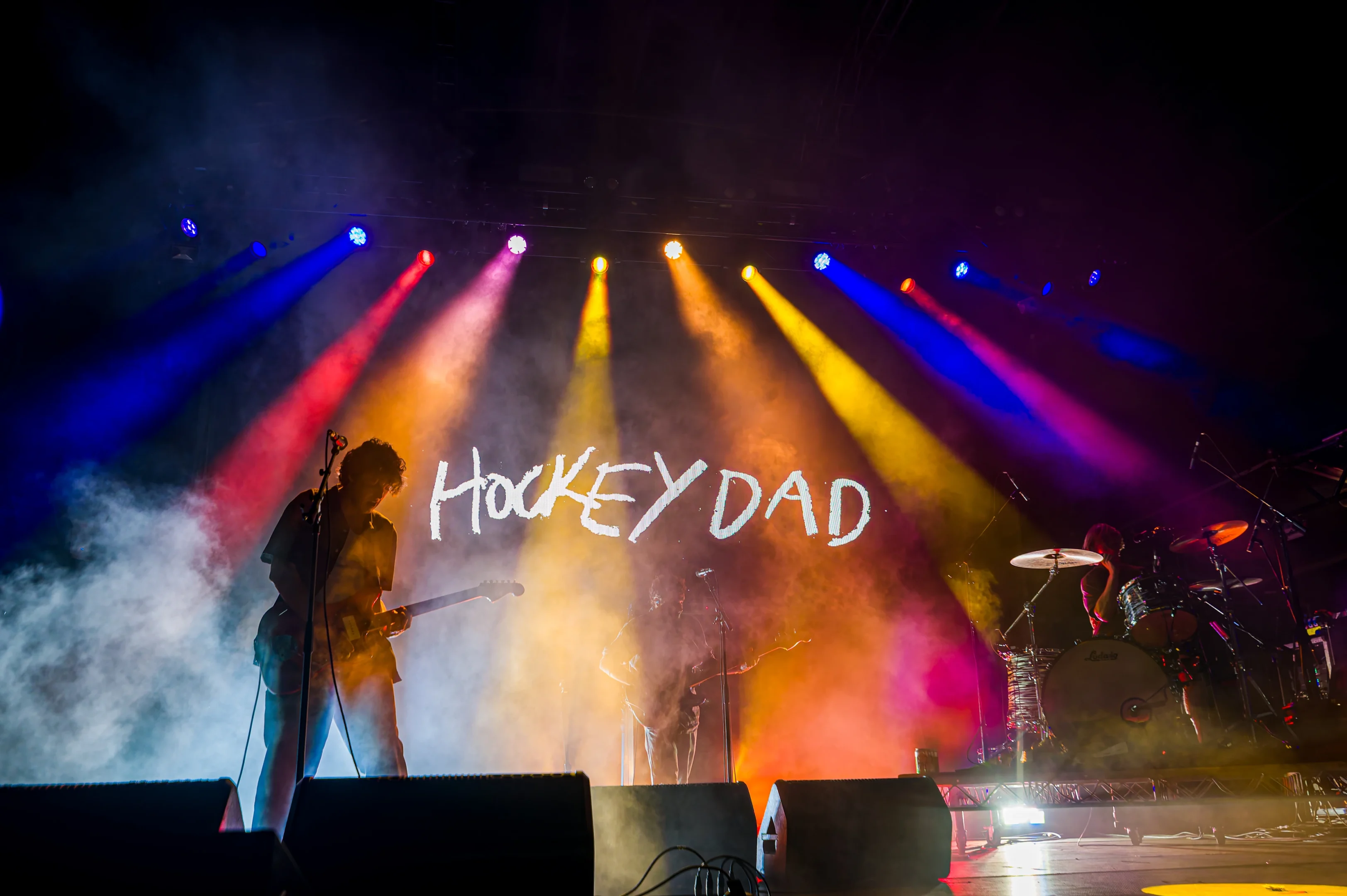 Band Performing With Colourful Lights, Smoke, And &Quot;Hockey Dad&Quot; Displayed In The Background.