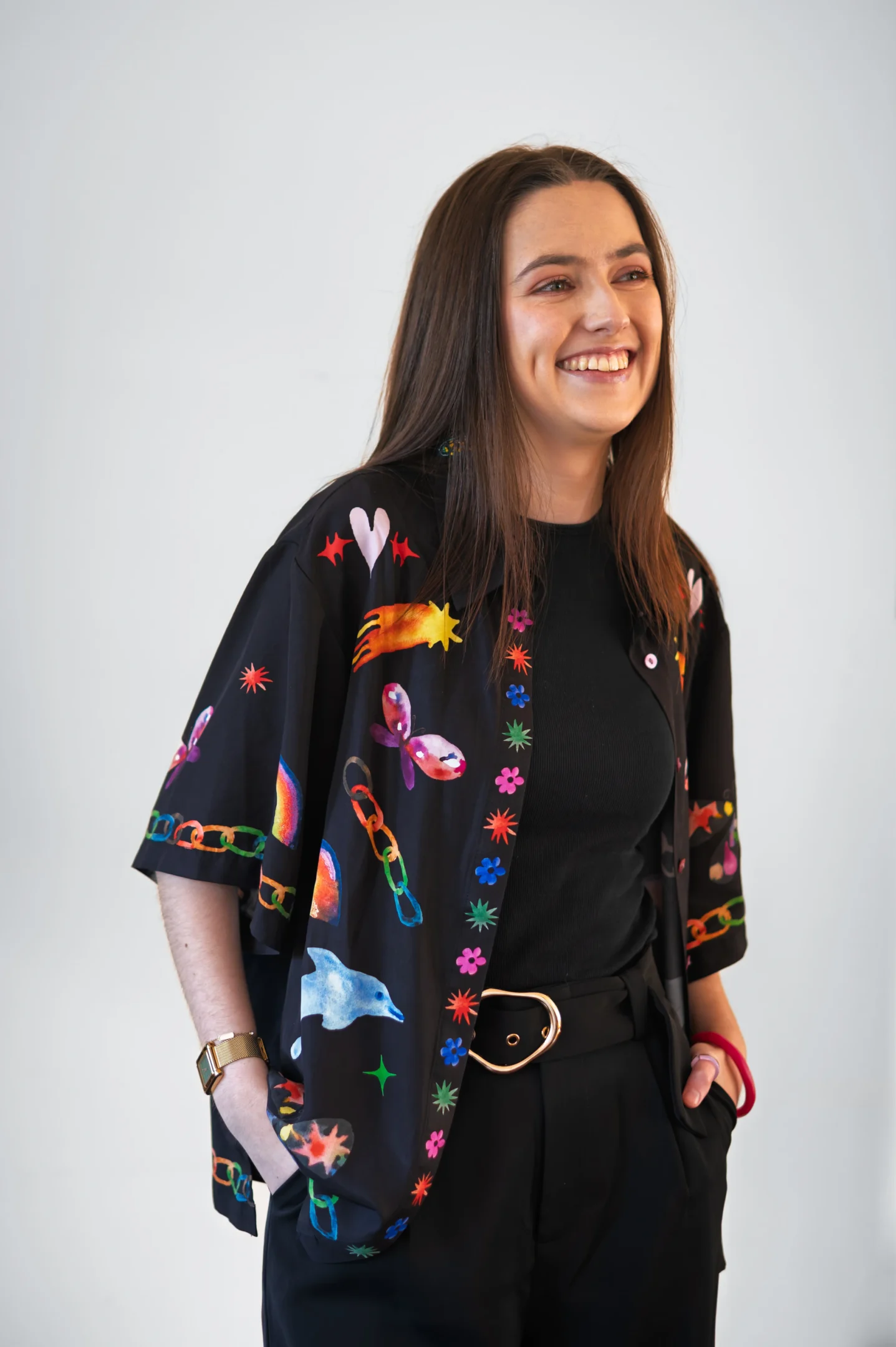 Person With Long Hair Smiles In Colourful Shirt, Black Top, Pants, Gold Watch, Circular Buckle.