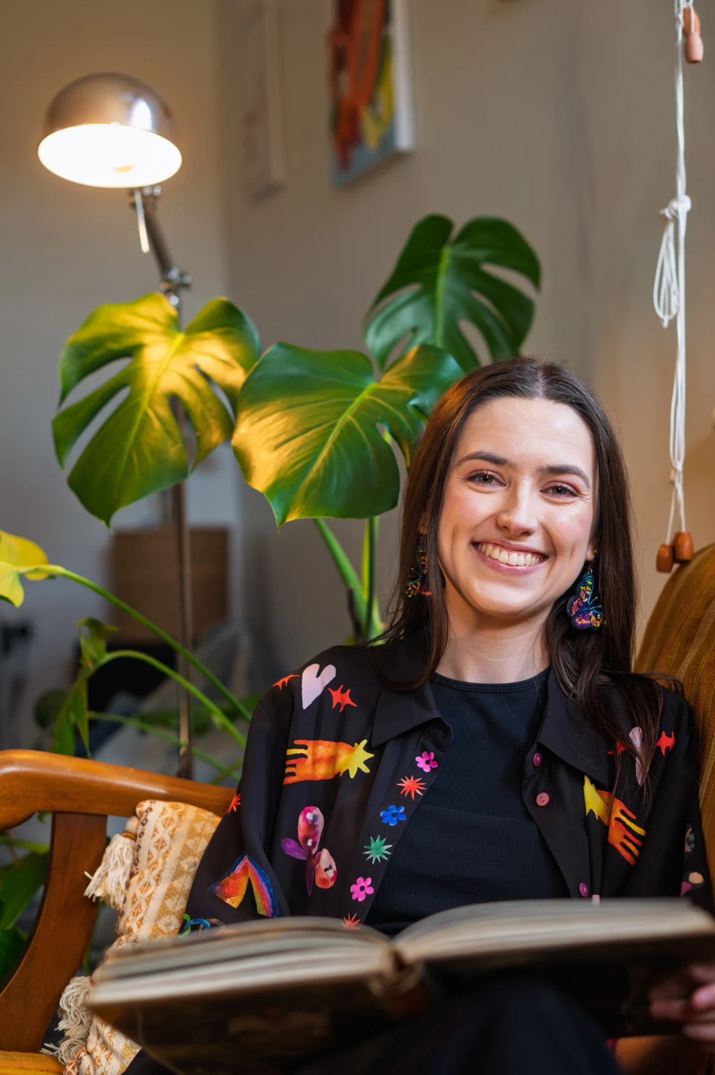 Person Seated Indoors, Smiling, In Colorful Shirt With Book, Green Plant, And Lamp Behind.