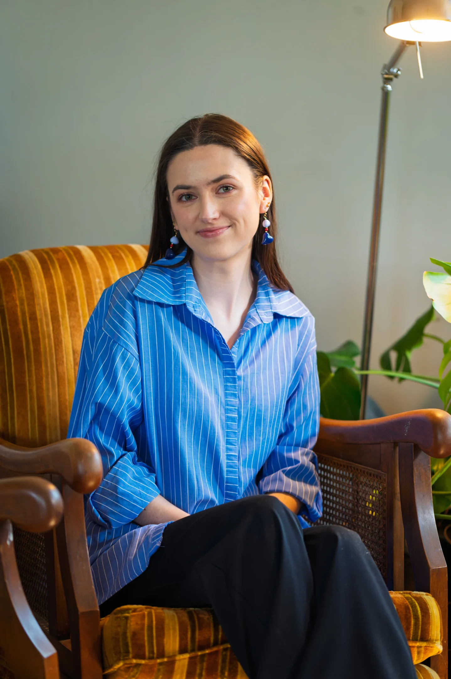 Person With Long Hair Seated On Brown Chair, Wearing A Blue Striped Shirt And Blue Earrings.