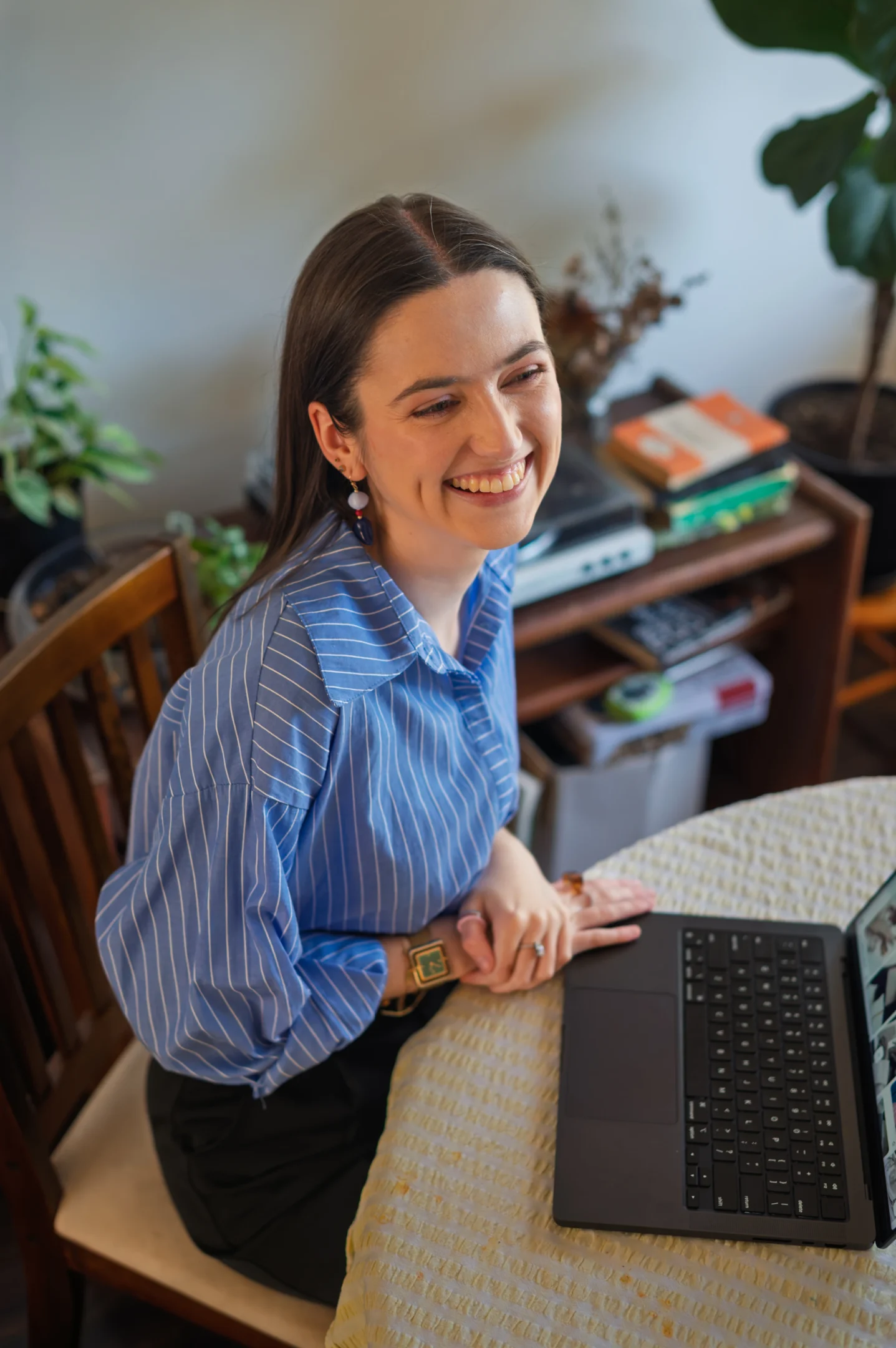 Woman In Blue Striped Shirt, Smiling At Laptop, With Plants And Bookshelf In Background.