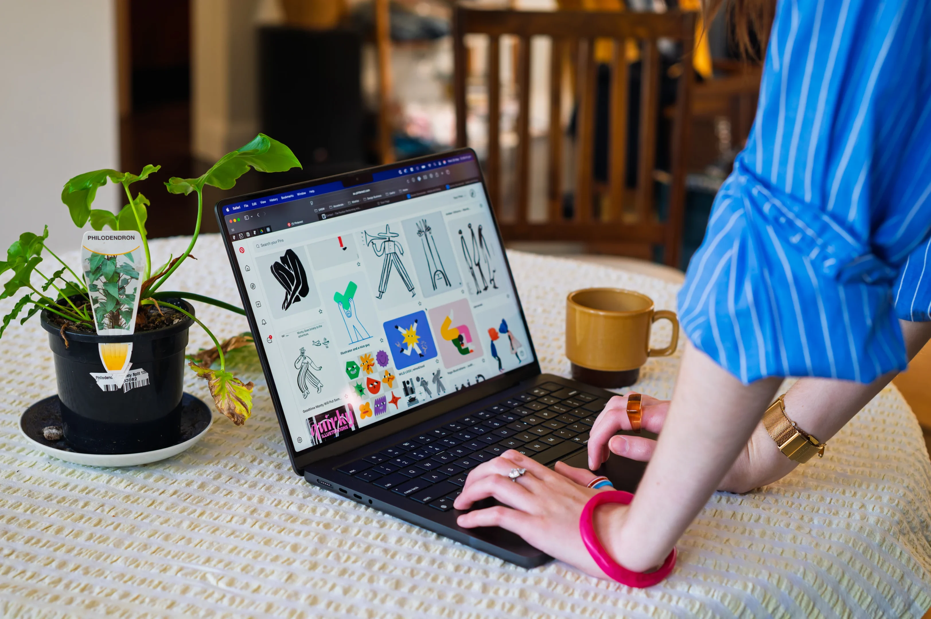 Person In BlueStriped Shirt Using Laptop With Graphic Designs; Plant And Mug On Table.