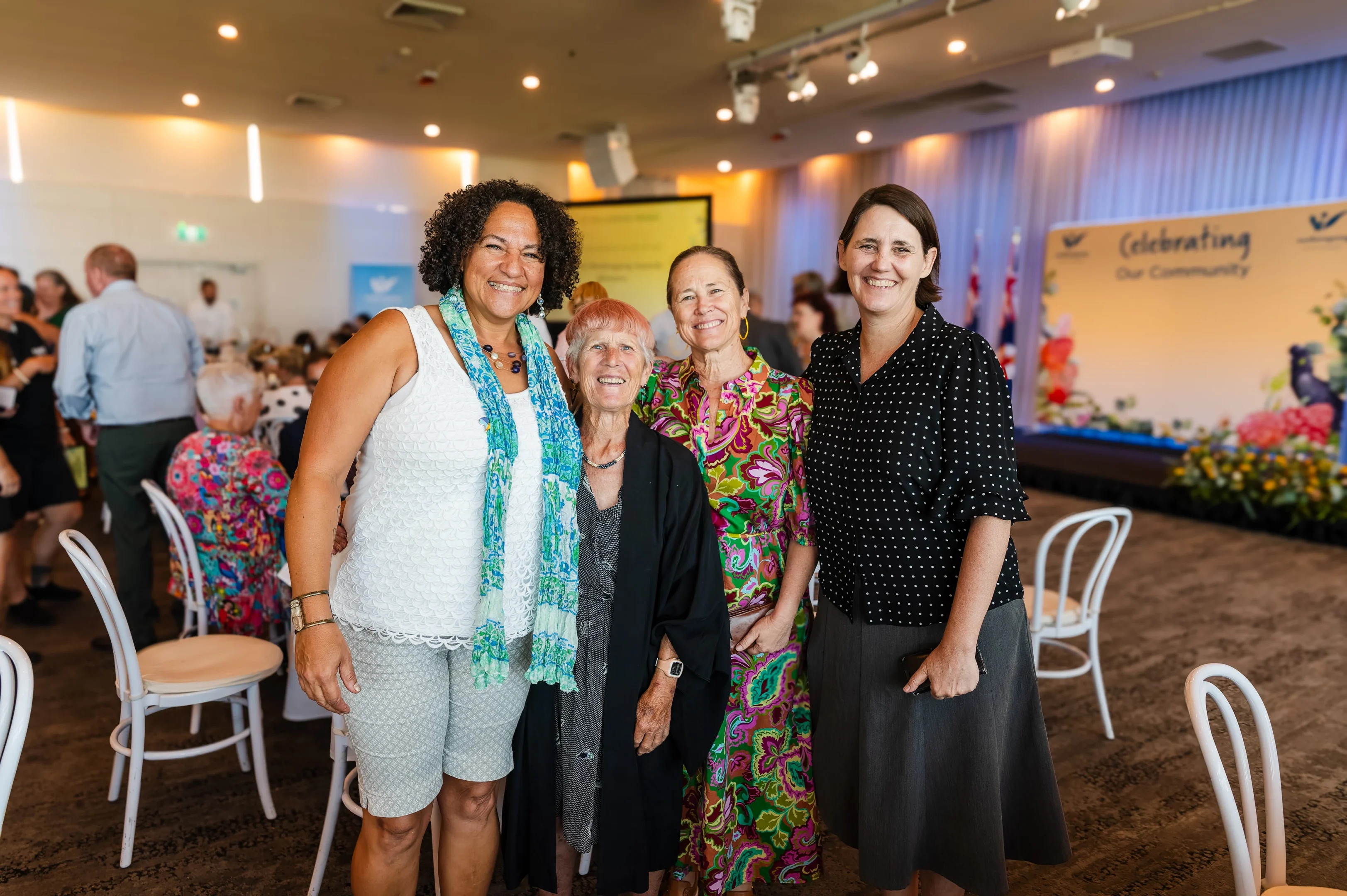Tips On Event Photography: Getting Comfortable, Capturing Moments, And Connecting With Guests 1 Four Women Smiling In A Festive Room With A &Quot;Celebrating Our Community&Quot; Sign And Floral Decor.